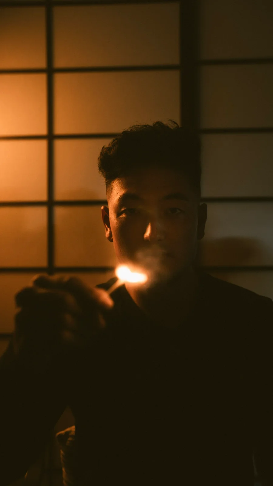 Harrison Saito in the dojo at night, lit by a single small flame, shoji glowing behind him