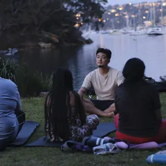 People seated together on the grass at dusk, the harbour lights behind