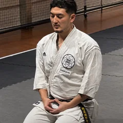 Harrison kneeling in seiza on the dojo mats in his gi and black belt, eyes closed